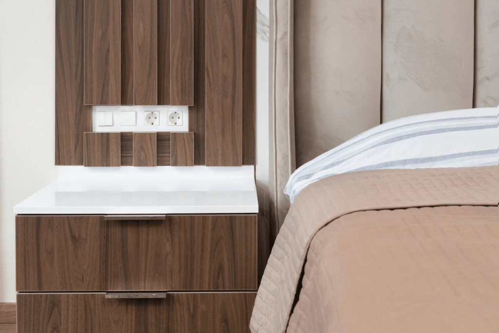 Ein Hotelbett mit einem Nachttisch und Steckdosen. Die Wand hinter dem Bett ist gepolster und Hinter dem Nachttisch befindet sich eine Holzwand aus dunklem Holz